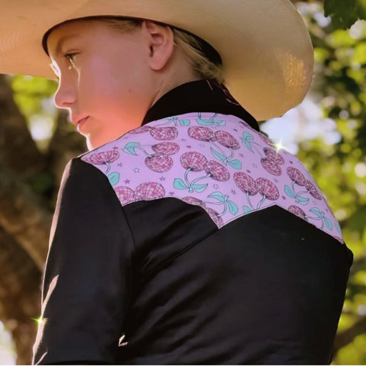 Young girl in a cowboy hat, and a black rodeo shirt with pink cherry accents showing the back of the shirt.