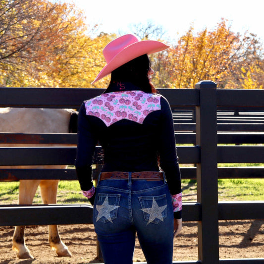 Woman in western attire with a pink hat and black rodeo shirt with cherry designs showing the back of the shirt, standing next to a saddle pad.