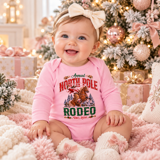 Smiling 6-month-old baby wearing a pink North Pole Rodeo Christmas romper featuring Cowboy Santa riding a bucking bronc, seated on a soft pink blanket in front of a blush pink and gold Christmas tree with wrapped presents, warm lights, and festive holiday decor.