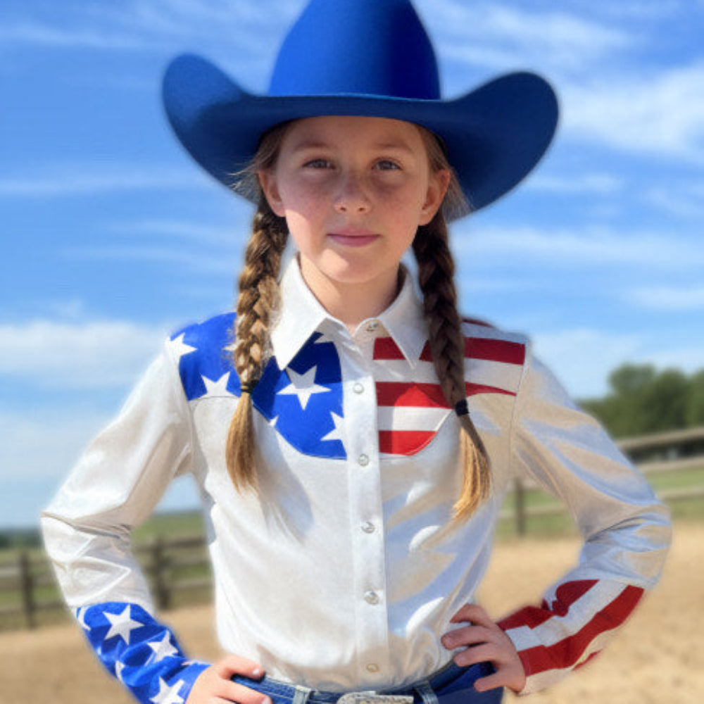 Young girl in a western rodeo shirt with an American flag design and blue hat in a dirt arena.