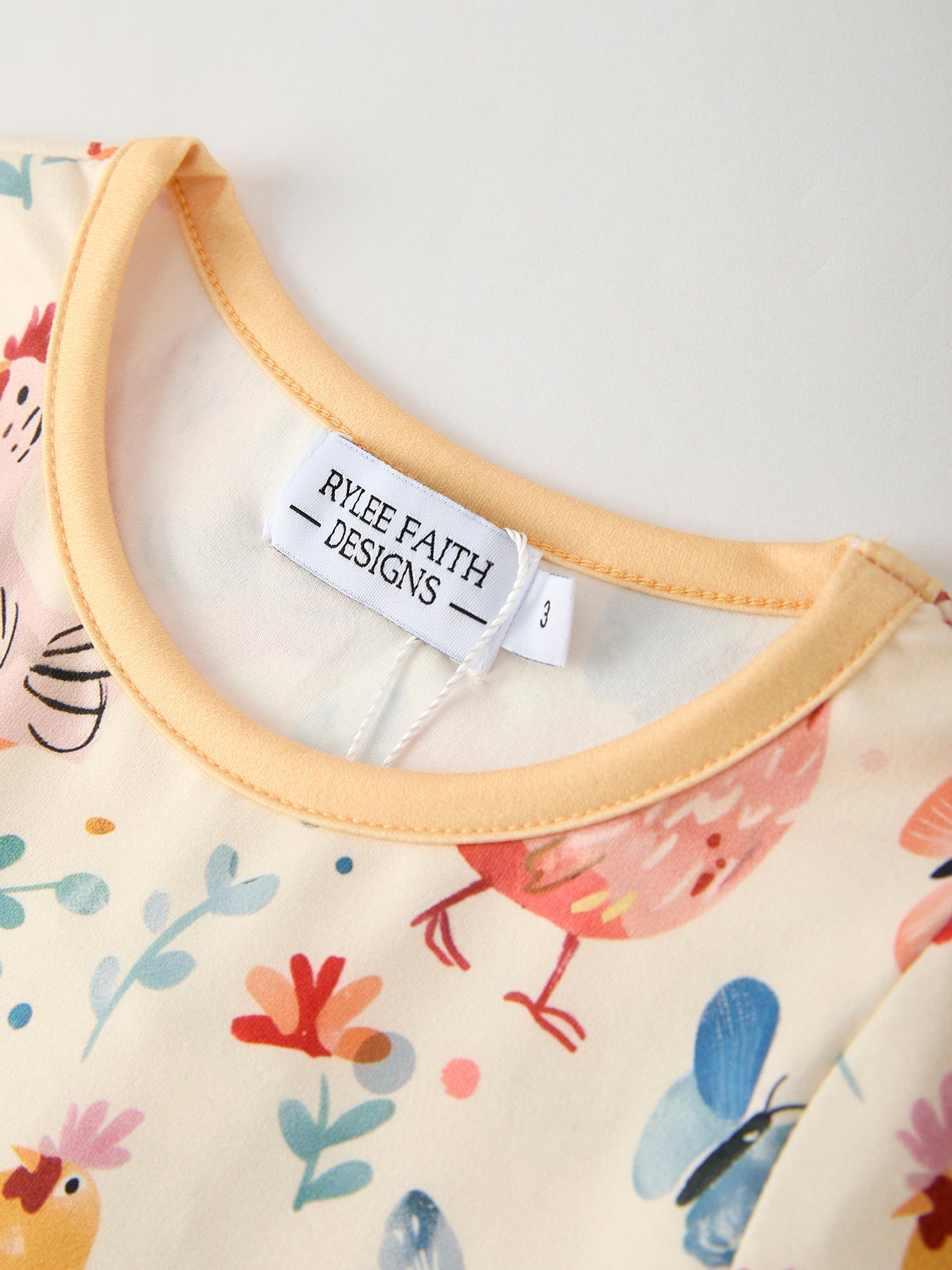 Close-up of a baby romper with colorful chicken and butterfly designs and 'Rylee Faith Designs' label.