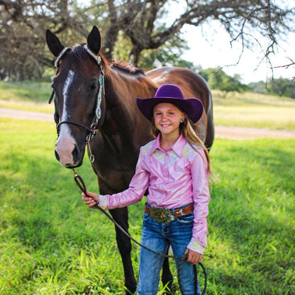 Young girl in a pink rodeo shirt and purple hat standing next to a brown horse in a grassy field.