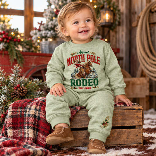 Smiling 12-month-old baby wearing a heather sage green North Pole Rodeo Christmas two-piece sweatsuit featuring Cowboy Santa riding a bucking bronc, paired with brown moccasins. Seated on a rustic wooden crate surrounded by festive plaid blankets, snowy pine trees, warm Christmas lights, and Western ranch decor.