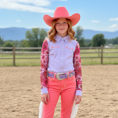 Young girl in pink salmon colored cowboy hat and pants standing in an outdoor setting with a fence and trees in the background.