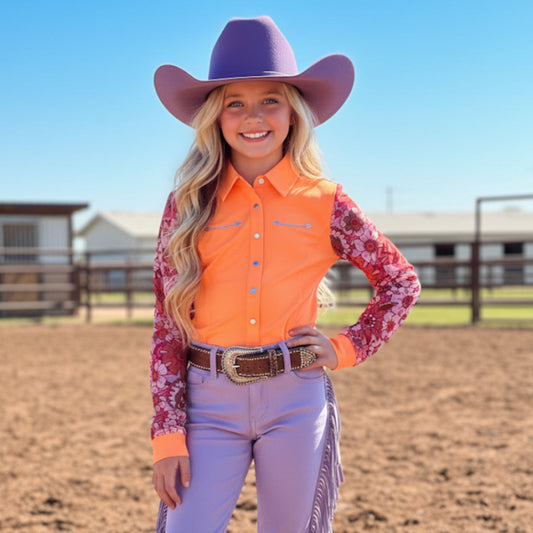 A girl wearing a retro pink and coral orange floral mesh sleeve rodeo shirt, purple pants, and a purple cowboy hat in a dirt area with buildings in the background.