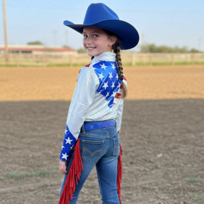 Young girl in a western rodeo shirt with an American flag design and blue hat in a dirt arena showing the back of the shirt.