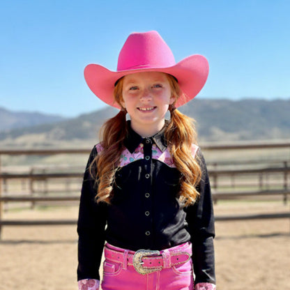 Young girl in a pink cowboy hat, a black rodeo shirt with pink cherry accents and pink pants standing in an outdoor setting with mountains in the background.
