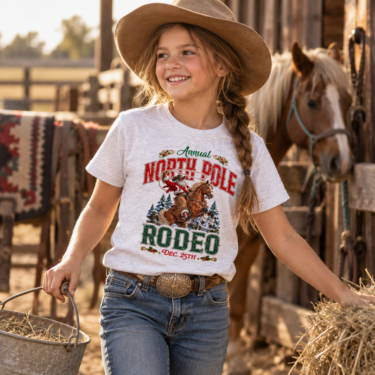 Smiling youth girl wearing a white North Pole Rodeo graphic T-shirt, blue jeans, a western belt, and a cowboy hat, carrying a metal bucket and working near hay bales in a rustic ranch setting. A horse stands in the background beside a barn, with warm golden light and vintage Western details creating a classic country scene.
