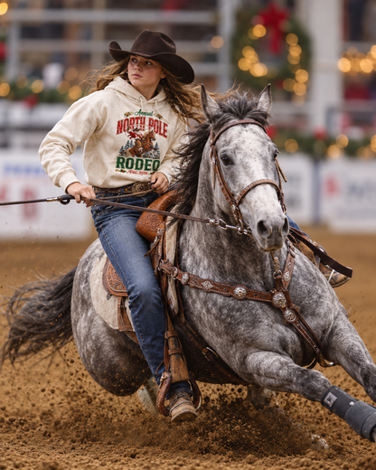 A young barrel racer in a cream “Annual North Pole Rodeo” hoodie and black cowboy hat rides a dapple gray horse in a sharp turn inside an arena. Dirt flies beneath the horse’s hooves, and soft Christmas lights and greenery blur in the background.