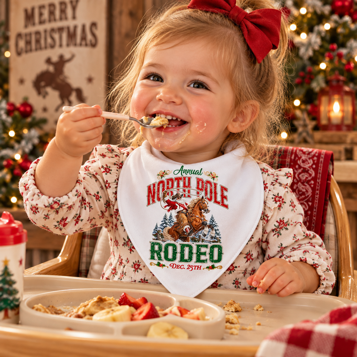 Smiling toddler girl sitting in a high chair eating food, wearing a white North Pole Rodeo Christmas bandana bib featuring Cowboy Santa riding a rearing horse. The scene has a cozy Western holiday theme with warm lights, festive decor, and Christmas trees in the background.