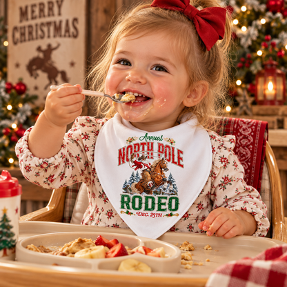 Smiling toddler girl sitting in a high chair eating food, wearing a white North Pole Rodeo Christmas bandana bib featuring Cowboy Santa riding a rearing horse. The scene has a cozy Western holiday theme with warm lights, festive decor, and Christmas trees in the background.