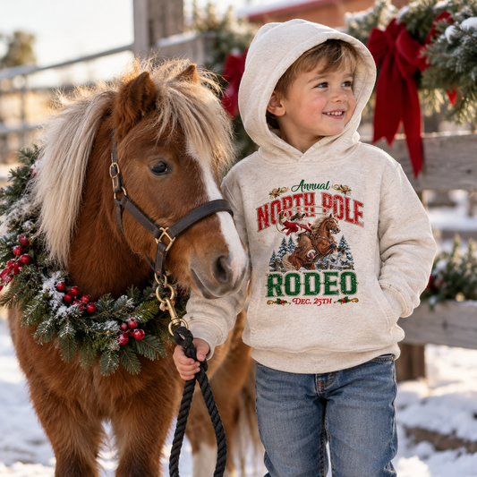 Smiling toddler boy wearing an oatmeal North Pole Rodeo Christmas hoodie with the hood up, blue jeans, and winter boots, standing in the snow beside a small pony decorated with festive greenery. The outdoor scene features a rustic fence, red bows, and a cozy Christmas ranch setting.