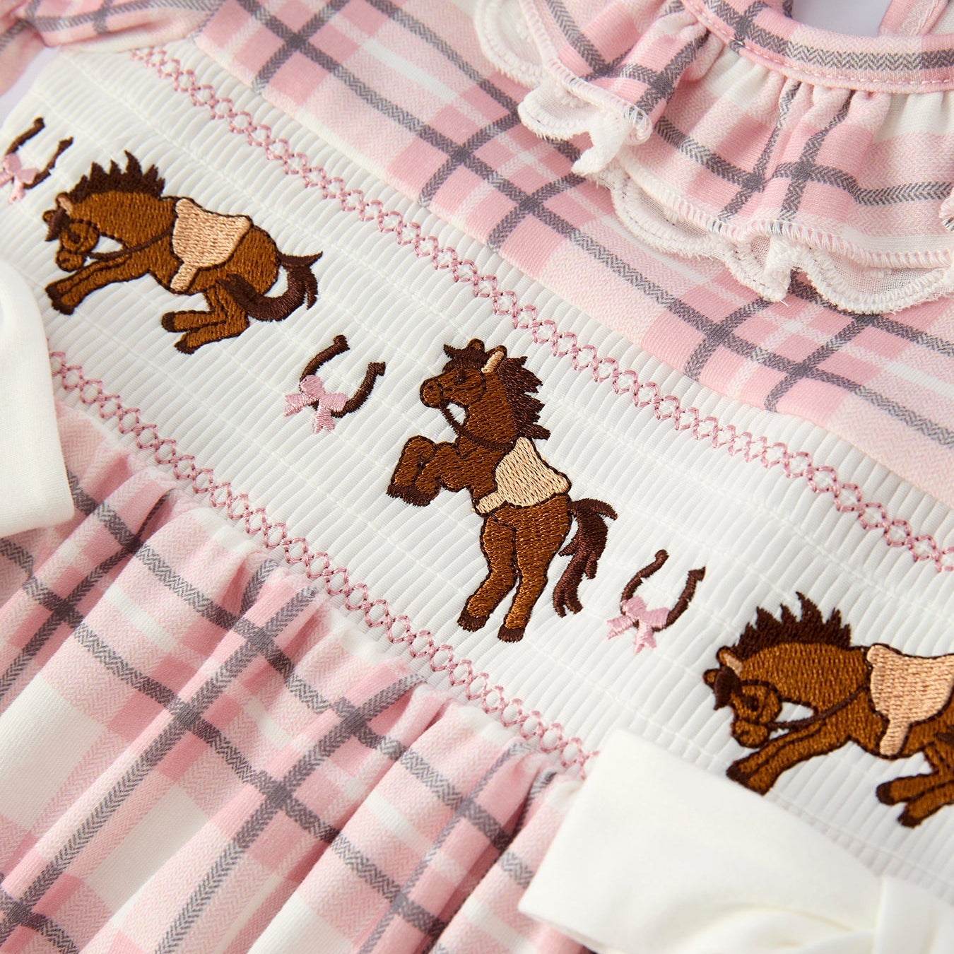 close up of embroidery on a pink plaid dress with horse design on a hanger against a white background