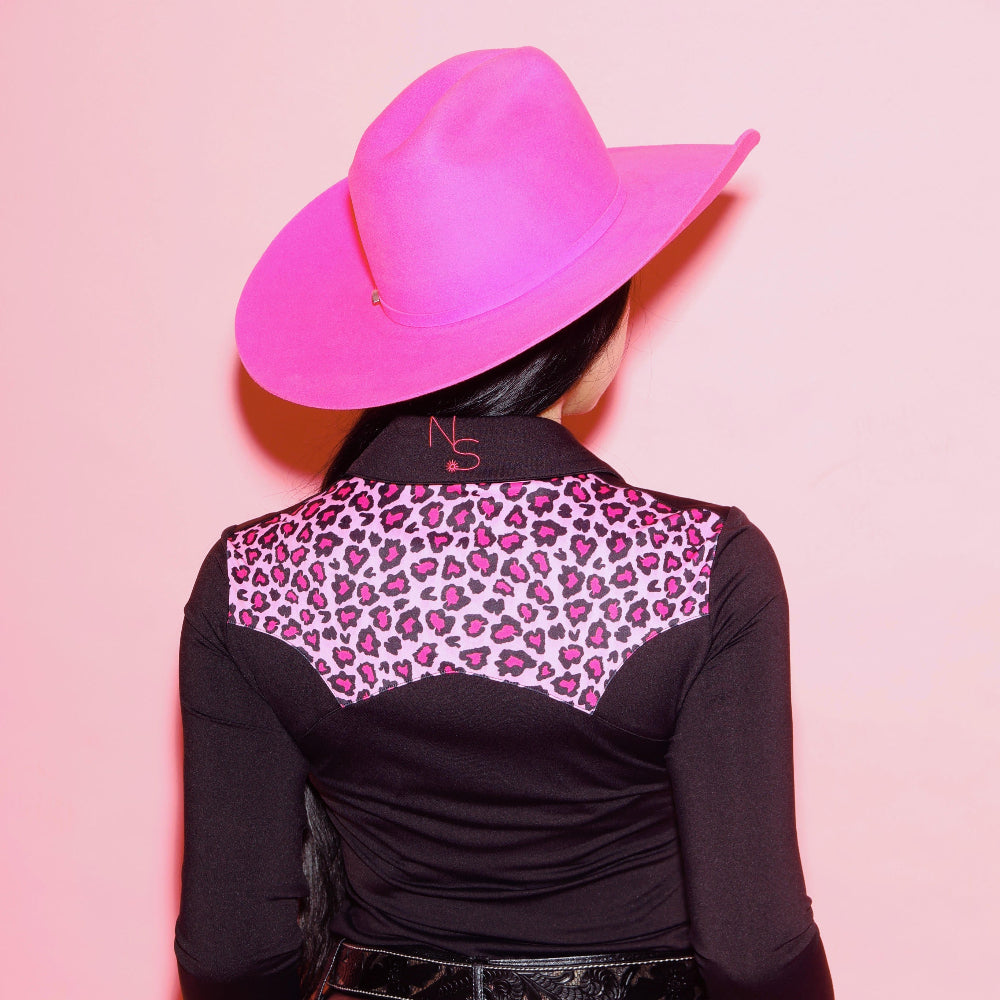 Woman wearing a black rodeo shirt with pink leopard print accents and a pink cowboy hat on a pink background showing the back of the shirt