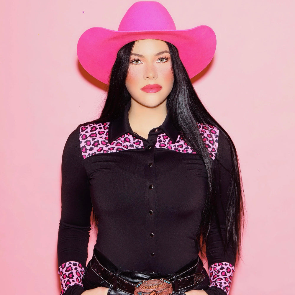 Woman wearing a black rodeo shirt with pink leopard print accents and a pink cowboy hat on a pink background
