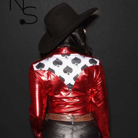 Woman wearing a red metallic rodeo pearl snap shirt with black spade accents and a black hat on a dark background showing the back of the shirt.