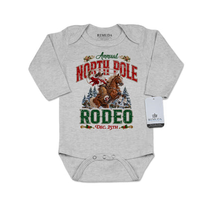 Heather Gray infant long sleeve Western Christmas romper featuring Cowboy Santa riding a rearing horse, festive North Pole Rodeo lettering in red and green, snowy pine trees, envelope-style shoulders, and snap closures for easy diaper changes.