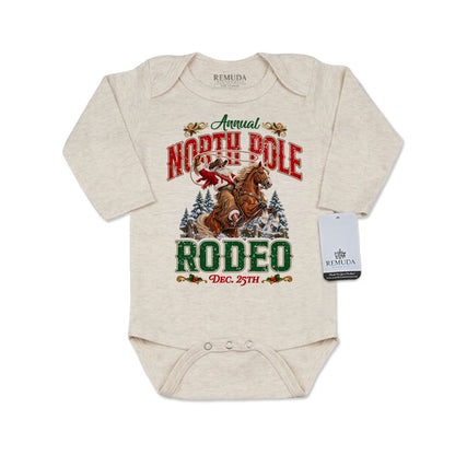 Oatmeal-colored infant long sleeve Western Christmas romper featuring Cowboy Santa riding a rearing horse, festive North Pole Rodeo lettering in red and green, snowy pine trees, envelope-style shoulders, and snap closures for easy diaper changes.