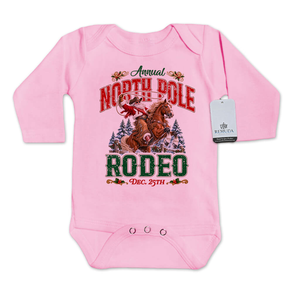Pink infant long sleeve Western Christmas romper featuring Cowboy Santa riding a rearing horse, festive North Pole Rodeo lettering in red and green, snowy pine trees, envelope-style shoulders, and snap closures for easy diaper changes.
