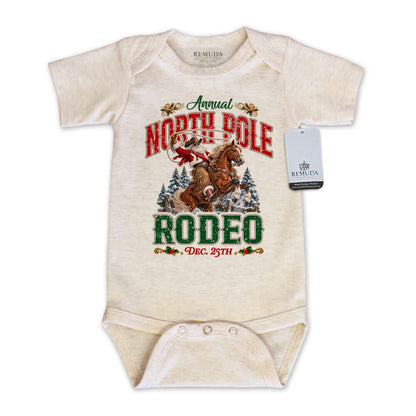 Oatmeal-colored infant short sleeve Western Christmas romper featuring Cowboy Santa riding a rearing horse, festive North Pole Rodeo lettering in red and green, snowy pine trees, envelope-style shoulders, and snap closures for easy diaper changes.