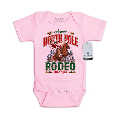 Pink infant short sleeve Western Christmas romper featuring Cowboy Santa riding a rearing horse, festive North Pole Rodeo lettering in red and green, snowy pine trees, envelope-style shoulders, and snap closures for easy diaper changes.