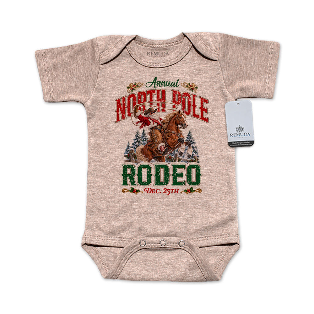 Sandstone-colored infant short sleeve Western Christmas romper featuring Cowboy Santa riding a rearing horse, festive North Pole Rodeo lettering in red and green, snowy pine trees, envelope-style shoulders, and snap closures for easy diaper changes.