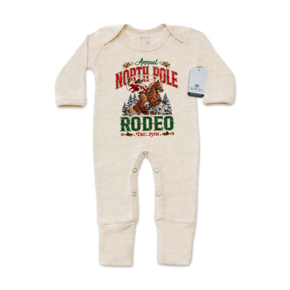 Oatmeal-colored infant one-piece footed pajamas featuring a festive North Pole Rodeo Christmas graphic with cowboy Santa riding a rearing horse, red and green lettering, long sleeves, and snap closures.