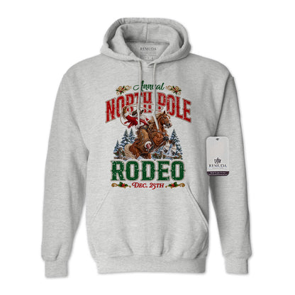 Ash Gray adult unisex pullover fleece hoodie featuring the Annual North Pole Rodeo Western Christmas design with Cowboy Santa riding a rearing horse, snowy pine trees, and festive red and green lettering reading “North Pole Rodeo – Dec. 25th.” Features a drawstring hood, kangaroo pocket, and ribbed cuffs and hem.