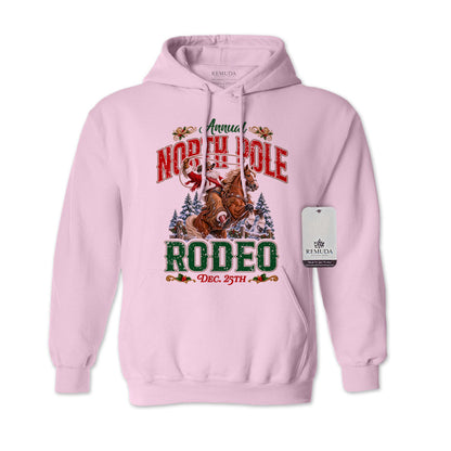 Light Pink adult unisex pullover fleece hoodie featuring the Annual North Pole Rodeo Western Christmas design with Cowboy Santa riding a rearing horse, snowy pine trees, and festive red and green lettering reading “North Pole Rodeo – Dec. 25th.” Features a drawstring hood, kangaroo pocket, and ribbed cuffs and hem.