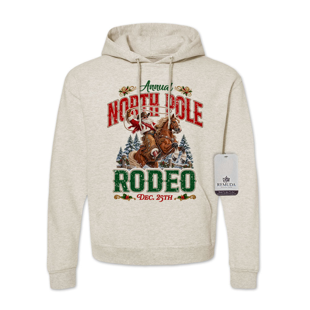 Oatmeal-colored adult unisex pullover fleece hoodie featuring the Annual North Pole Rodeo Western Christmas design with Cowboy Santa riding a rearing horse, snowy pine trees, and festive red and green lettering reading “North Pole Rodeo – Dec. 25th.” Features a drawstring hood, kangaroo pocket, and ribbed cuffs and hem.