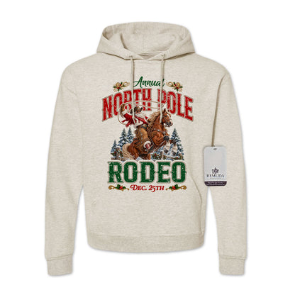 Oatmeal-colored adult unisex pullover fleece hoodie featuring the Annual North Pole Rodeo Western Christmas design with Cowboy Santa riding a rearing horse, snowy pine trees, and festive red and green lettering reading “North Pole Rodeo – Dec. 25th.” Features a drawstring hood, kangaroo pocket, and ribbed cuffs and hem.