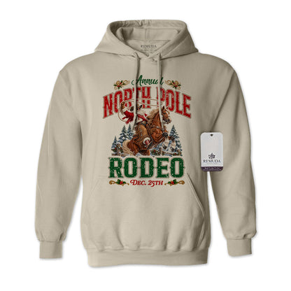 Sand-colored adult unisex pullover fleece hoodie featuring the Annual North Pole Rodeo Western Christmas design with Cowboy Santa riding a rearing horse, snowy pine trees, and festive red and green lettering reading “North Pole Rodeo – Dec. 25th.” Features a drawstring hood, kangaroo pocket, and ribbed cuffs and hem.