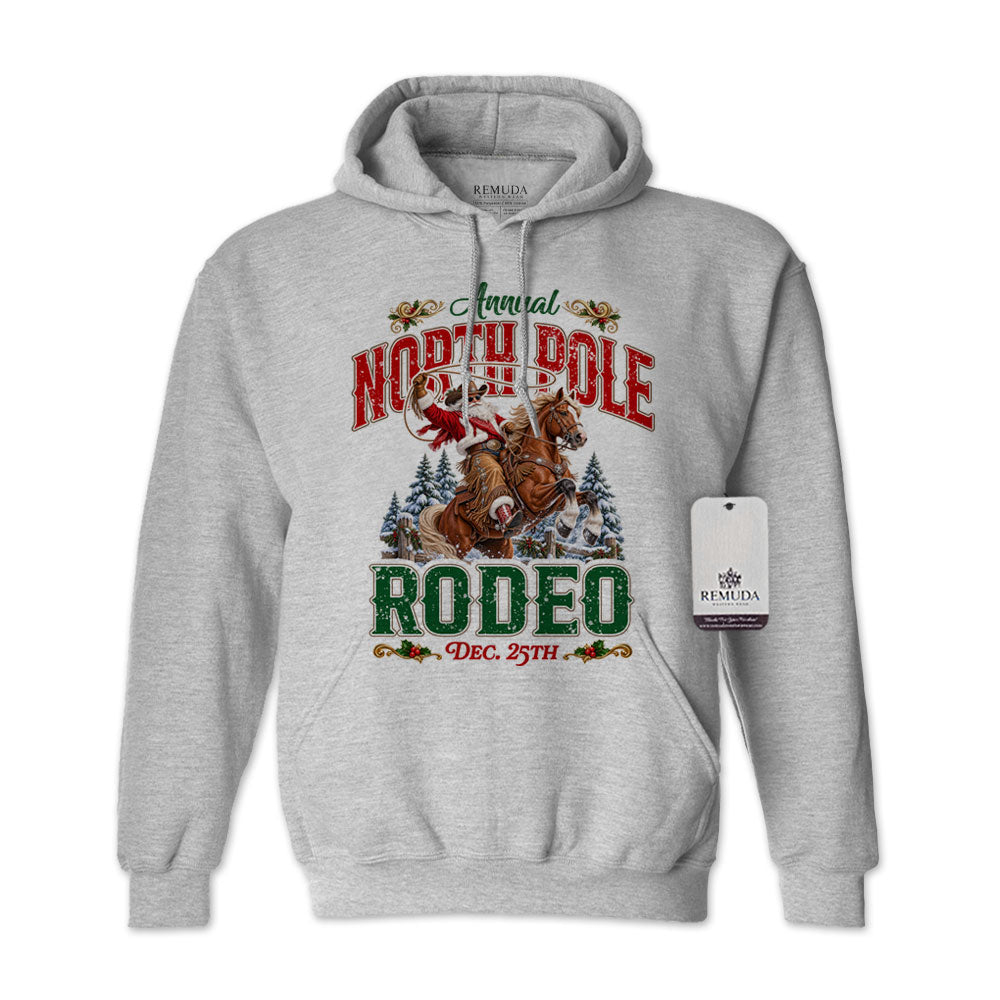 Sport Gray adult unisex pullover fleece hoodie featuring the Annual North Pole Rodeo Western Christmas design with Cowboy Santa riding a rearing horse, snowy pine trees, and festive red and green lettering reading “North Pole Rodeo – Dec. 25th.” Features a drawstring hood, kangaroo pocket, and ribbed cuffs and hem.