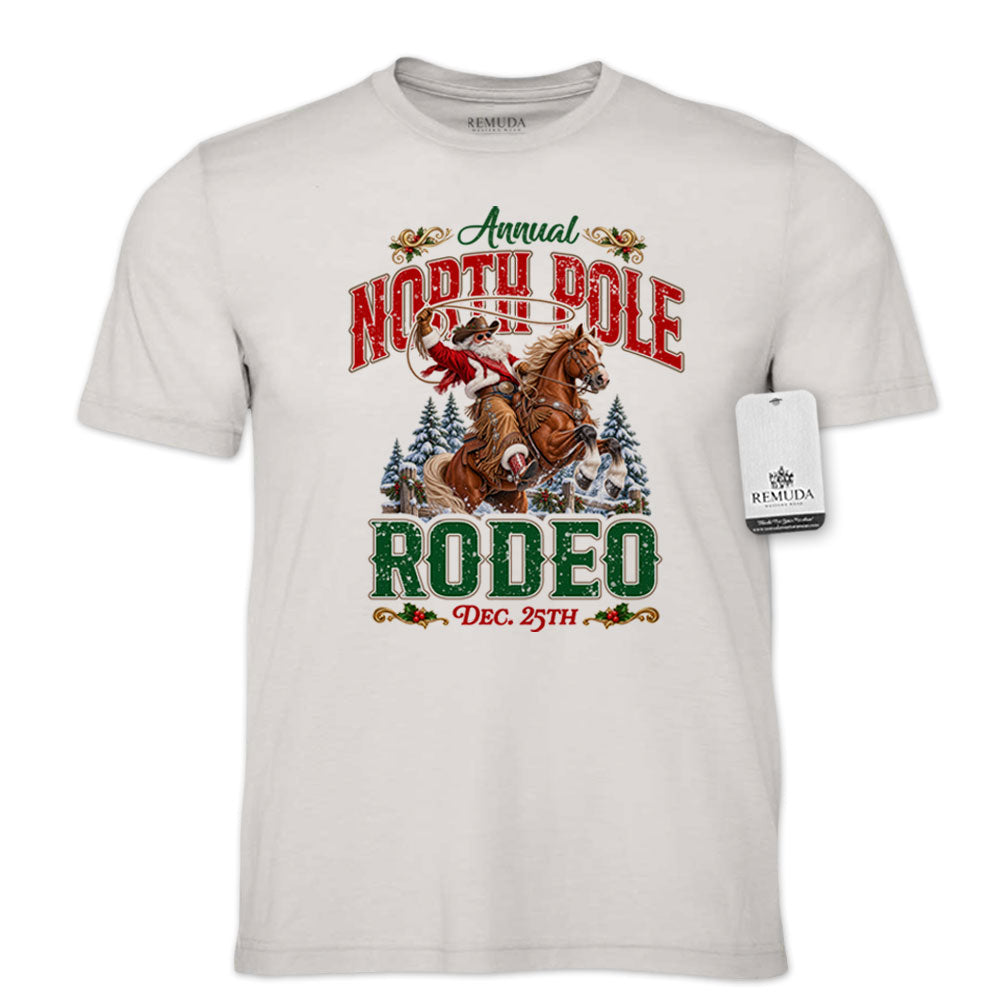 Cement-color adult unisex short sleeve T-shirt featuring a festive North Pole Rodeo Christmas design with Cowboy Santa riding a rearing horse, snowy pine trees, vintage red and green Western lettering, December 25th details, and a classic crewneck style.