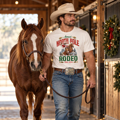 A cowboy wearing a cream North Pole Rodeo Christmas T-shirt, blue jeans, western hat, and turquoise-accented championship team roping belt buckle leads a large chestnut horse with a white blaze through a rustic barn aisle decorated with Christmas wreaths, greenery, and warm holiday lights.