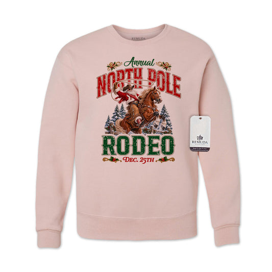 Blush pink adult unisex crewneck fleece sweatshirt featuring a festive North Pole Rodeo Christmas design with Cowboy Santa riding a rearing horse, snowy pine trees, vintage red and green Western lettering, December 25th details, and ribbed cuffs and hem.