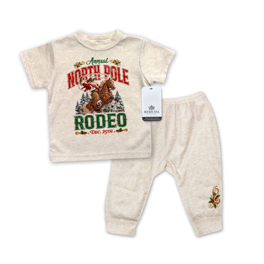 Oatmeal-colored infant two-piece Western Christmas pajama set featuring a short sleeve shirt with Cowboy Santa riding a rearing horse, festive red and green North Pole Rodeo lettering, and matching elastic-waist pants with decorative holiday embroidery.