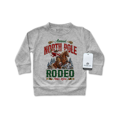 Heather gray infant long sleeve pullover sweatshirt featuring a festive North Pole Rodeo Christmas design with Cowboy Santa riding a rearing horse, snowy pine trees, vintage red and green lettering, and ribbed cuffs and hem.