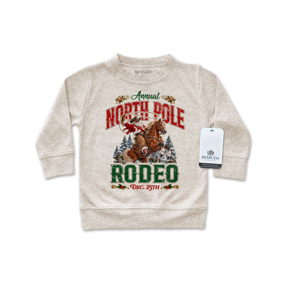 Oatmeal-colored infant long sleeve pullover sweatshirt featuring a festive North Pole Rodeo Christmas design with Cowboy Santa riding a rearing horse, snowy pine trees, vintage red and green lettering, and ribbed cuffs and hem.