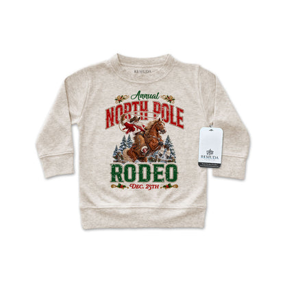 Oatmeal-colored infant long sleeve pullover sweatshirt featuring a festive North Pole Rodeo Christmas design with Cowboy Santa riding a rearing horse, snowy pine trees, vintage red and green lettering, and ribbed cuffs and hem.