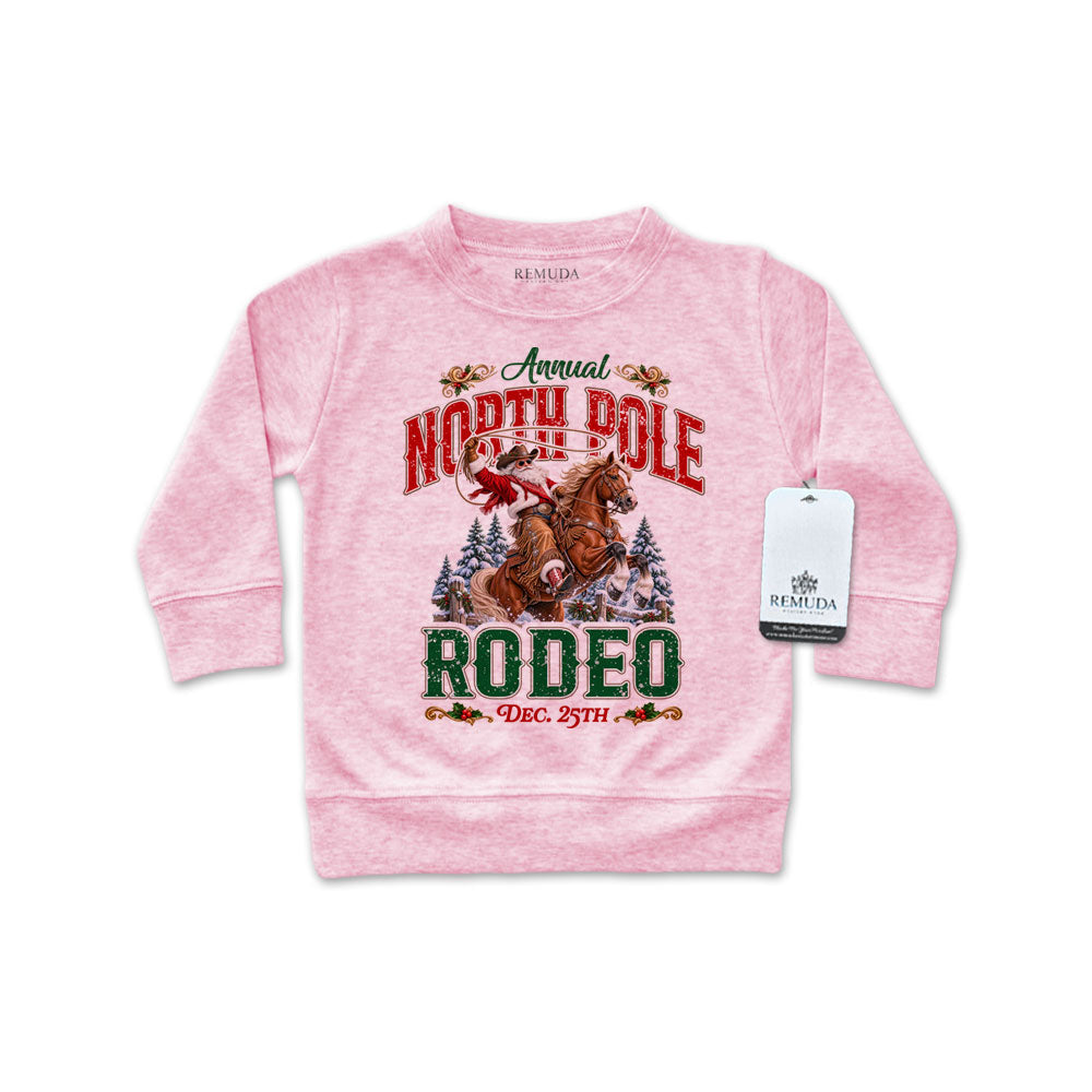 Pink infant long sleeve pullover sweatshirt featuring a festive North Pole Rodeo Christmas design with Cowboy Santa riding a rearing horse, snowy pine trees, vintage red and green lettering, and ribbed cuffs and hem.