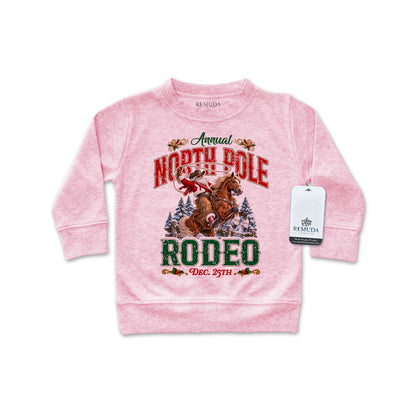 Pink infant long sleeve pullover sweatshirt featuring a festive North Pole Rodeo Christmas design with Cowboy Santa riding a rearing horse, snowy pine trees, vintage red and green lettering, and ribbed cuffs and hem.