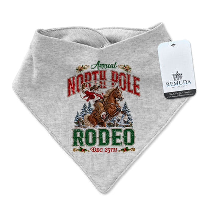 Heather Gray infant/toddler bandana bib featuring a festive North Pole Rodeo Christmas design with Cowboy Santa riding a rearing horse, red and green vintage lettering, snap closures at the neck, and a Remuda Western Wear product tag attached.