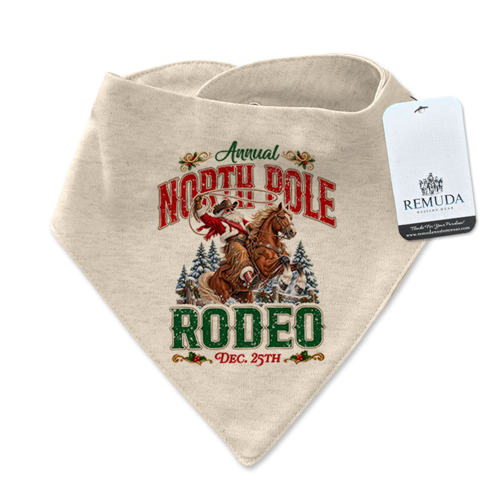 Oatmeal-colored infant/toddler bandana bib featuring a festive North Pole Rodeo Christmas design with Cowboy Santa riding a rearing horse, red and green vintage lettering, snap closures at the neck, and a Remuda Western Wear product tag attached.