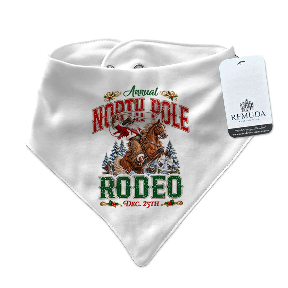 White infant/toddler bandana bib featuring a festive North Pole Rodeo Christmas design with Cowboy Santa riding a rearing horse, red and green vintage lettering, snap closures at the neck, and a Remuda Western Wear product tag attached.