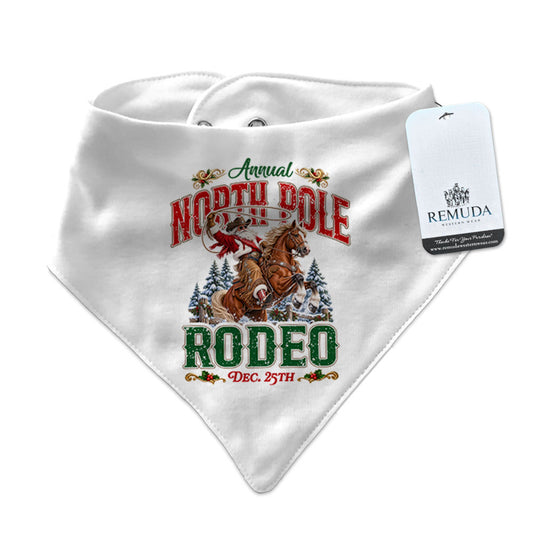 White infant/toddler bandana bib featuring a festive North Pole Rodeo Christmas design with Cowboy Santa riding a rearing horse, red and green vintage lettering, snap closures at the neck, and a Remuda Western Wear product tag attached.