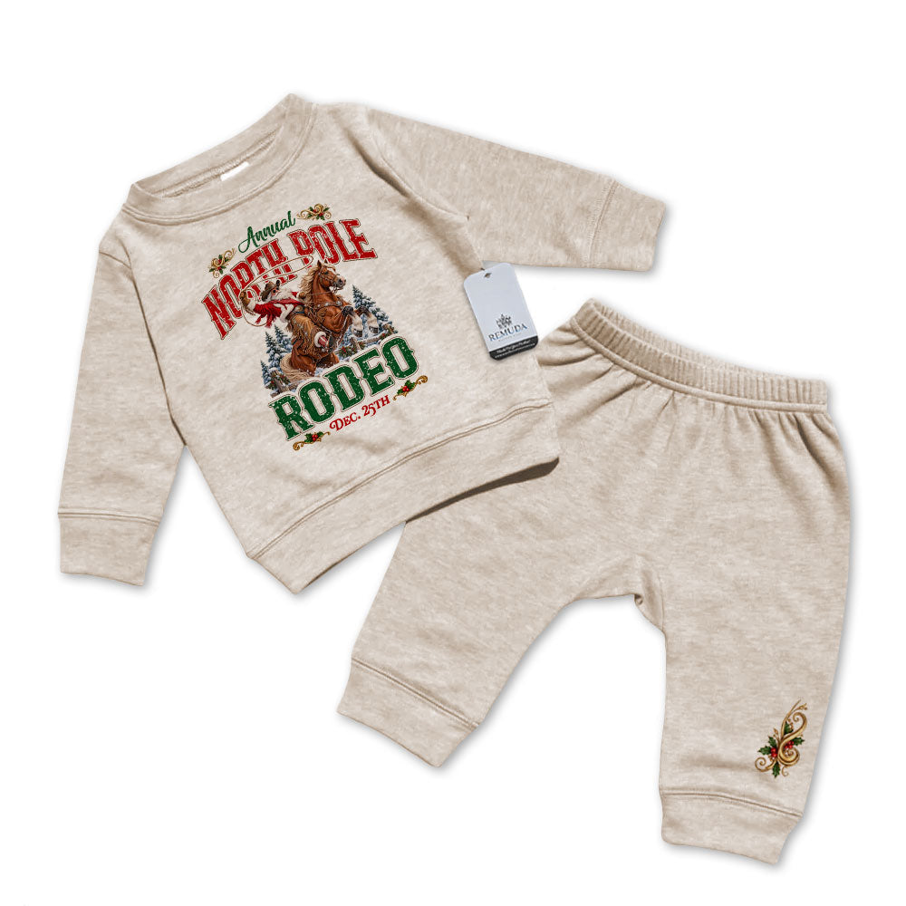 Oatmeal-colored infant two-piece sweatsuit featuring a long sleeve pullover sweatshirt with Cowboy Santa riding a bucking bronc in a festive North Pole Rodeo Christmas design, paired with matching elastic-waist sweatpants with a decorative holiday design.