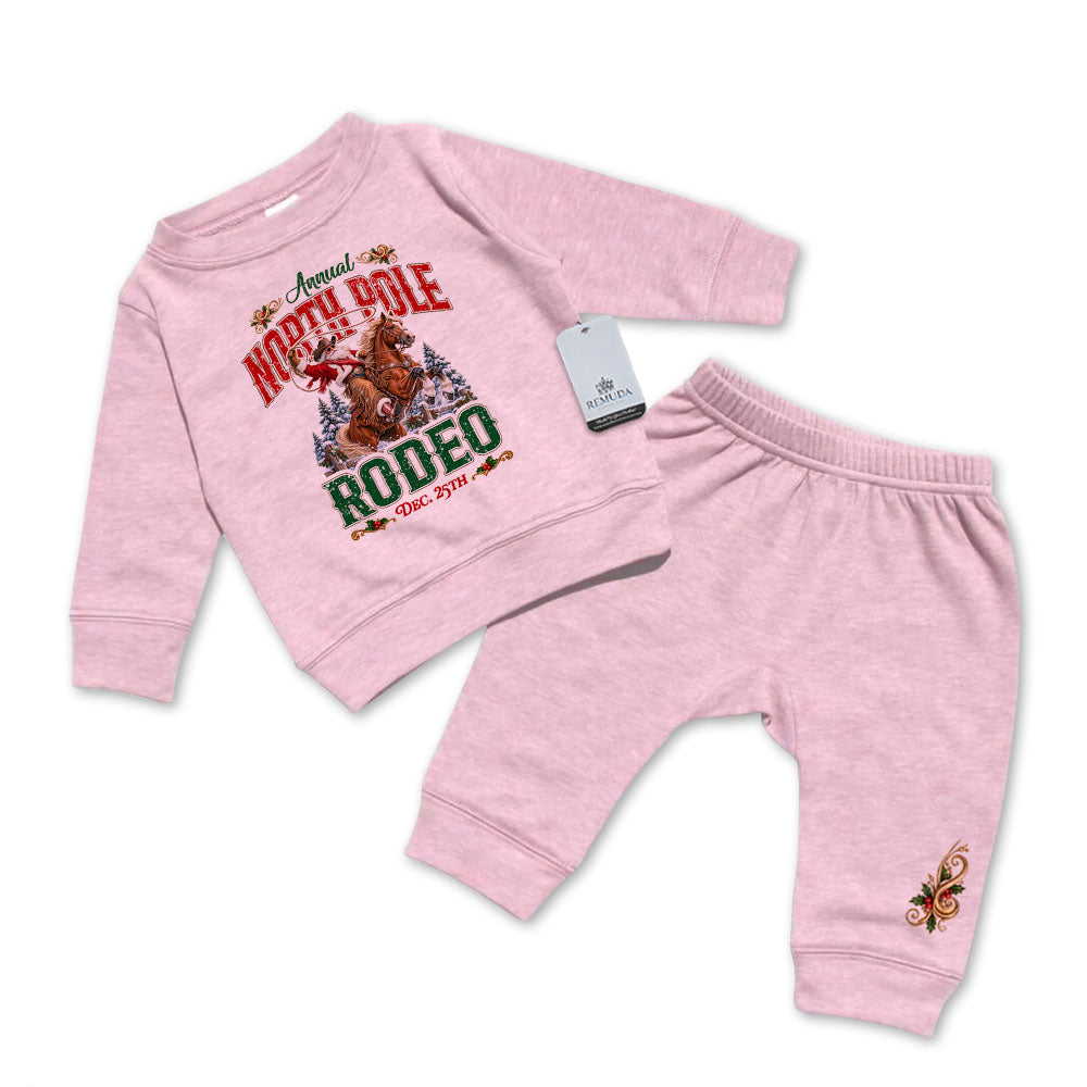 Pink-colored infant two-piece sweatsuit featuring a long sleeve pullover sweatshirt with Cowboy Santa riding a bucking bronc in a festive North Pole Rodeo Christmas design, paired with matching elastic-waist sweatpants with a decorative holiday design.