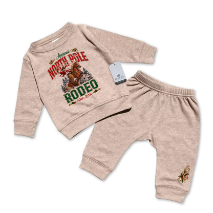 Sandstone-colored infant two-piece sweatsuit featuring a long sleeve pullover sweatshirt with Cowboy Santa riding a bucking bronc in a festive North Pole Rodeo Christmas design, paired with matching elastic-waist sweatpants with a decorative holiday design.
