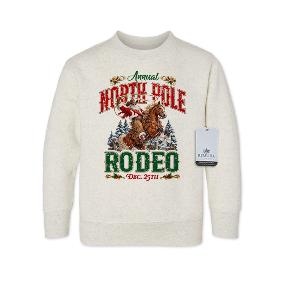 Oatmeal-colored toddler crewneck fleece sweatshirt featuring a festive North Pole Rodeo Christmas design with Cowboy Santa riding a rearing horse, snowy pine trees, vintage red and green lettering, December 25th details, and ribbed cuffs and hem.